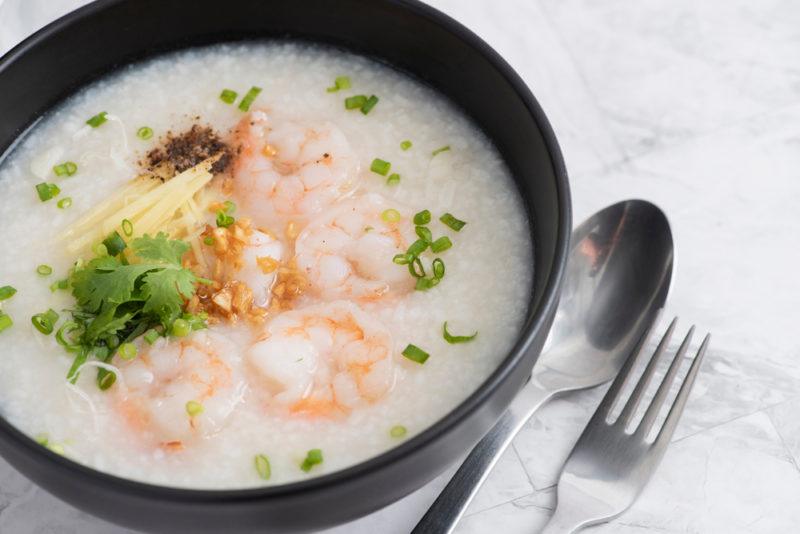 A black bowl of khao tom porridge with toppings, including shrimp, next to a fork and a spoon