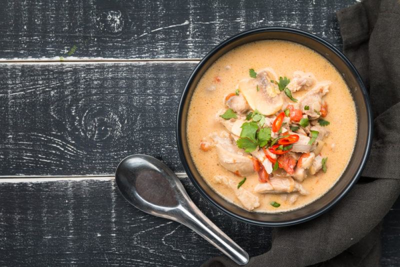 A black bowl with creamy tom kha gai on a black wooden table next to a spoon