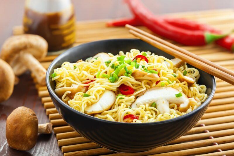 A black bowl filled with ramen noodles, some vegetables, and mushrooms, next to chopsticks and ingredients