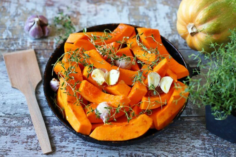 A black bowl filled with roasted pumpkin and garlic, next to herbs, a wooden spatula, and pumpkin