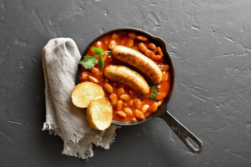 A cast iron pan filled with baked beans, with sausages and two slices of bread on top