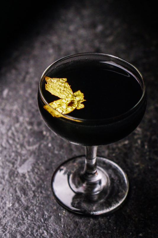 A black cocktail that uses a few gold leaves as a garnish