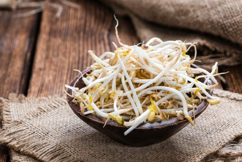 A wooden table with a woven cloth and a small black dish containing many mung bean sprouts.