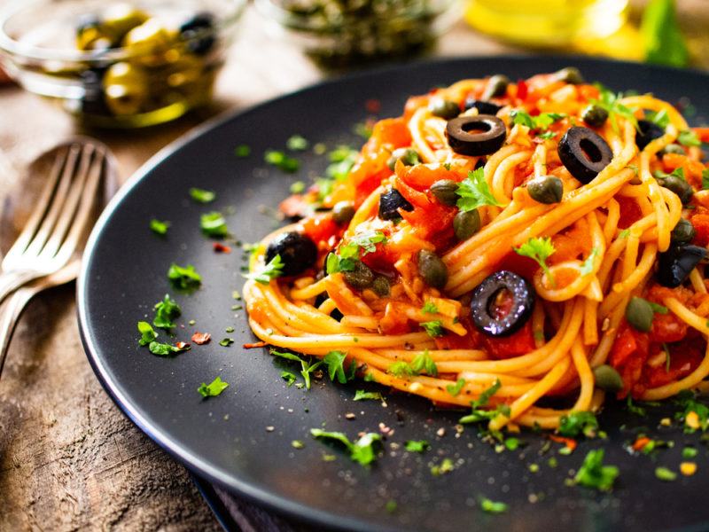 A black plate on a table with a dish of spaghetti puttanesca