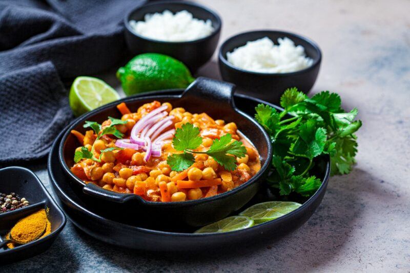 A black bowl with a bright red chickpea curry, next to some limes and smaller black bowls of rice