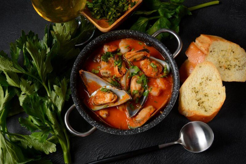 A black dish with handles, containing a tomato-based mussels meal, next to leafy greens and bread