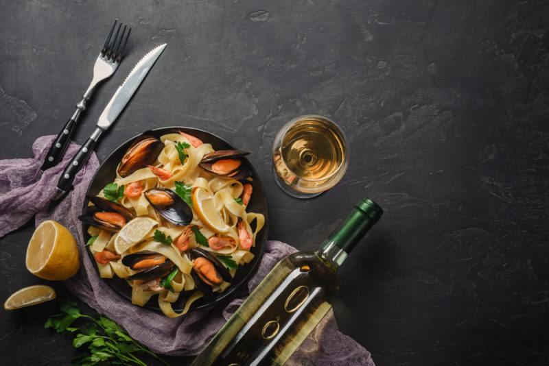 A black dish of pasta with cutlery on a table next to a glass of white wine