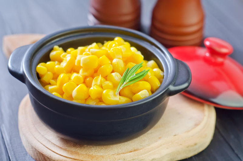 A small black dish filled with cooked sweet corn, next to a red lid for the dish and a salt and pepper grinder