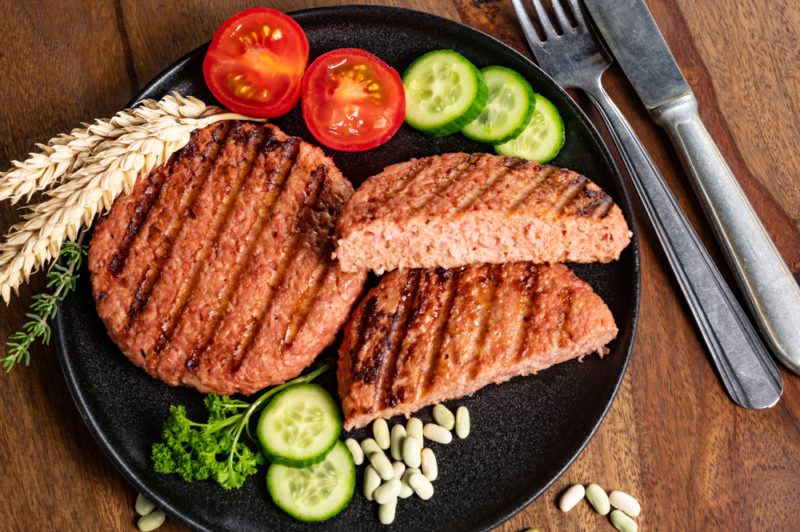 A black dish with two vegan burger patties, one of which has been cut in half. There is also some tomatoes and sliced cucumber