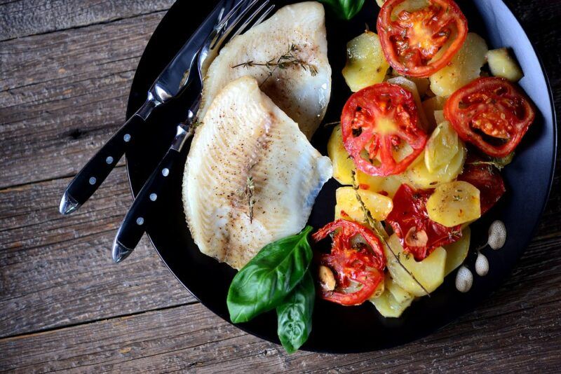 A black plate with cooked tilapia, potatoes, and tomatoes or peppers