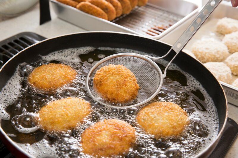 A frypan being used to deep fry croquettes, where one is being scooped out. There is a container of cooked croquettes and one of uncooked croquettes in the background.