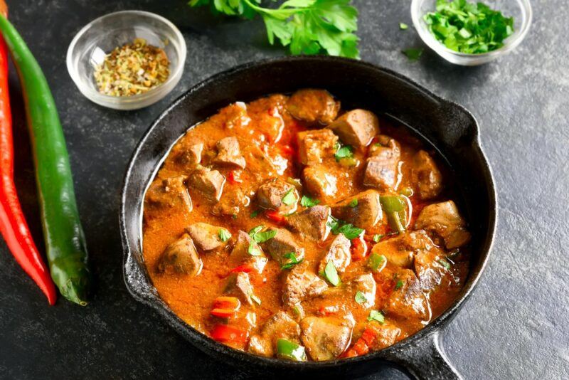 A black pan that contains a cooked chicken liver curry, next to a few hot peppers and other ingredients