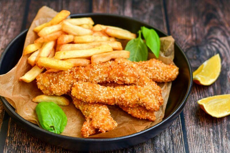 A black frypan containing a piece of greaseproof paper, fries, fried chicken, and green leaves, next to two lemon wedges