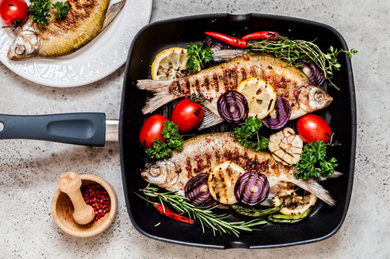 A black pan with two cooked fish, greens and tomatoes