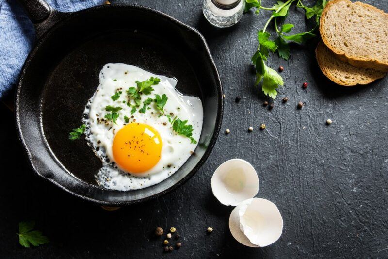 A black table with a cast iron frypan that contains a single fried egg. The egg shells are on the table, along with some pieces of bread.