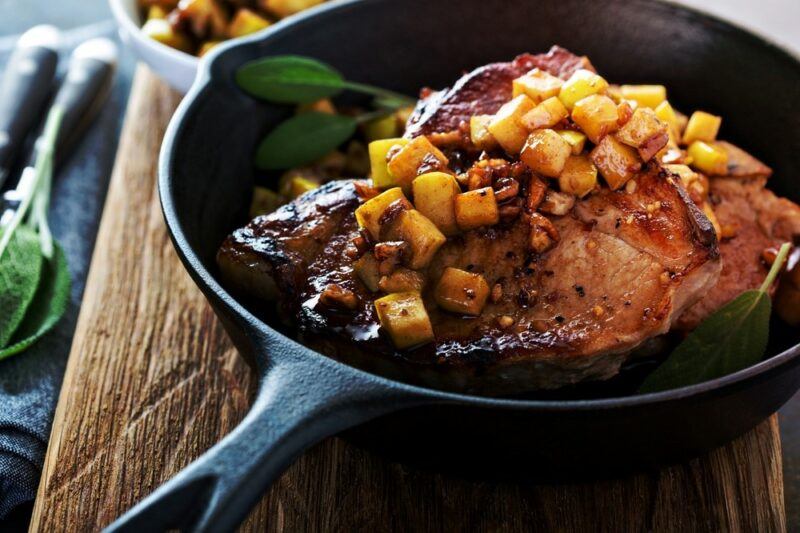 A wooden table with a black pan that contains a large piece of pork that has been covered in apples