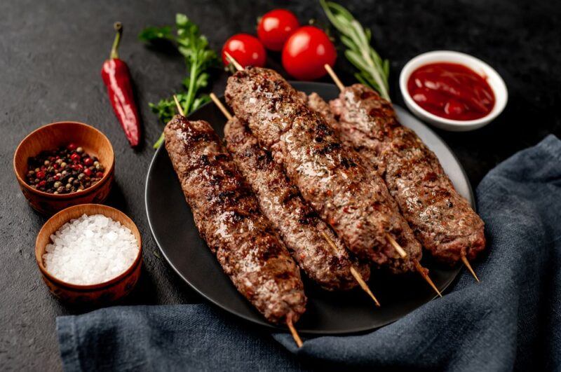 A black plate with five skewers of meat, surrounded by bowls of pepper, salt, and sauce, plus fresh herbs and tomatoes