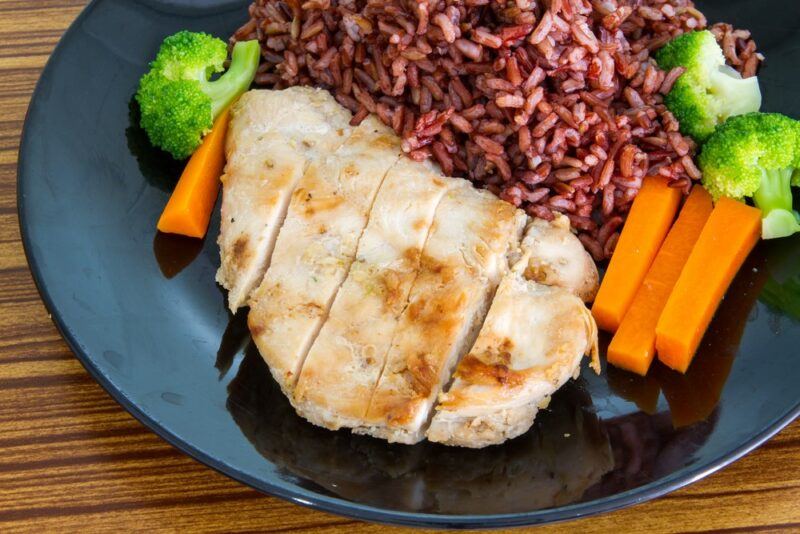 A black dish with chicken, brown rice, and some veggies