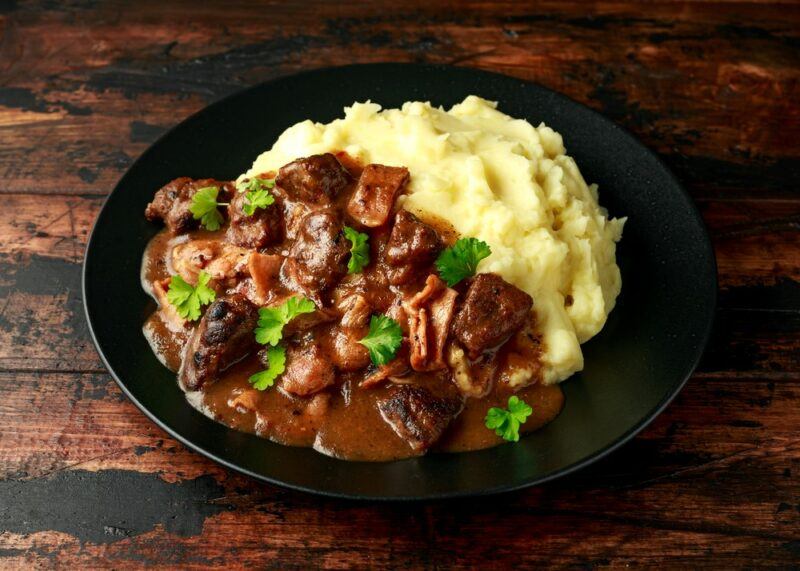 A black dish with a cooked liver stew and potatoes on a wooden table