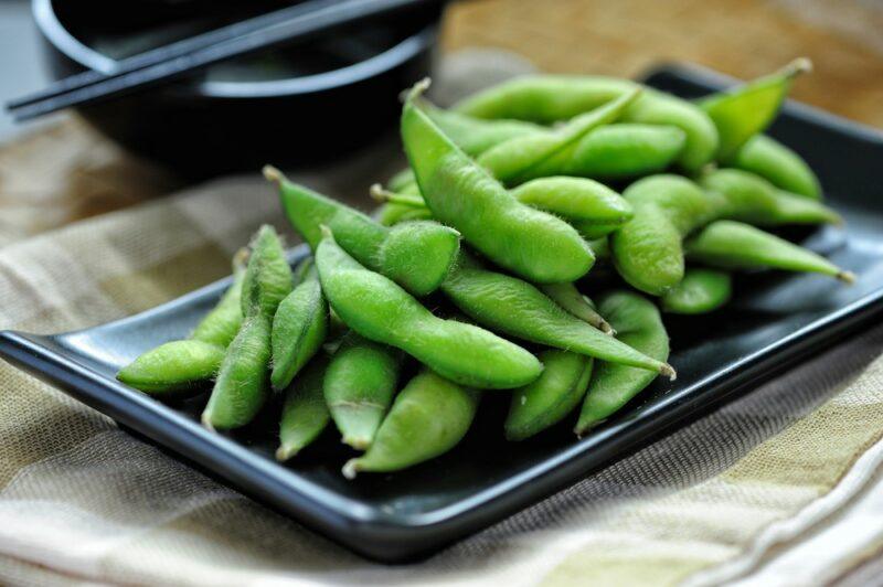 A black plate with edamame, next to a bowl that may contain dip or oil