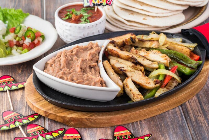 A wooden board with a black dish of fajita chicken, next to a dish of refried beans