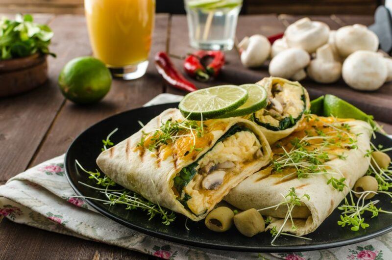 A black plate with a few homemade breakfast burritos with lime on top, with various ingredients in the background, including mushrooms and chili peppers