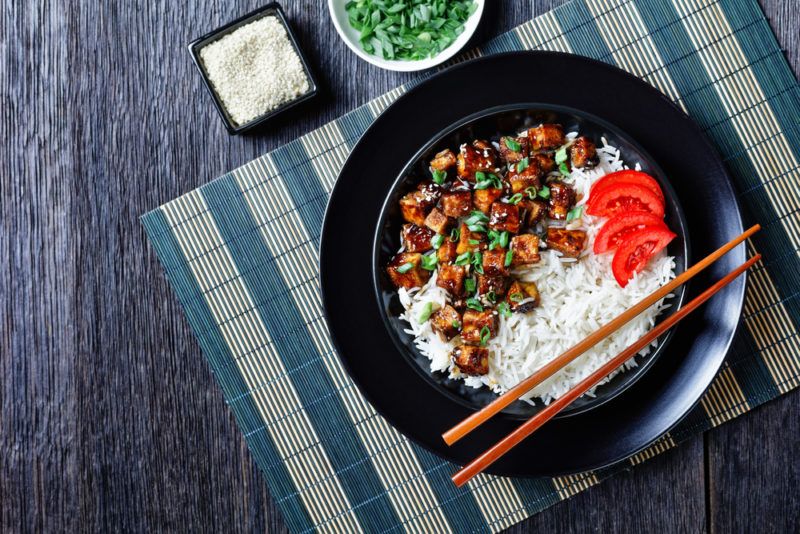 A black plate with white rice, chopsticks, and tofu, on a table cloth next to some seasoning ingredients