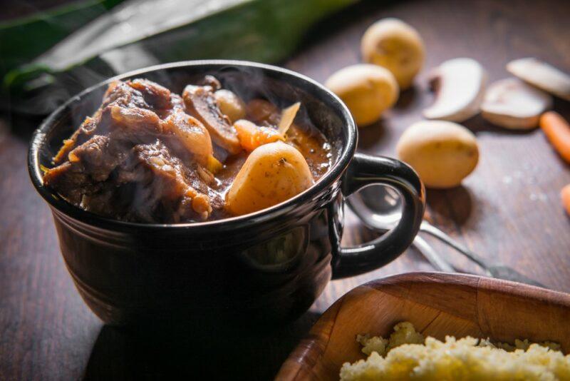 A black bowl or mug of oxtail soup on a table with some potatoes and mushrooms in the background