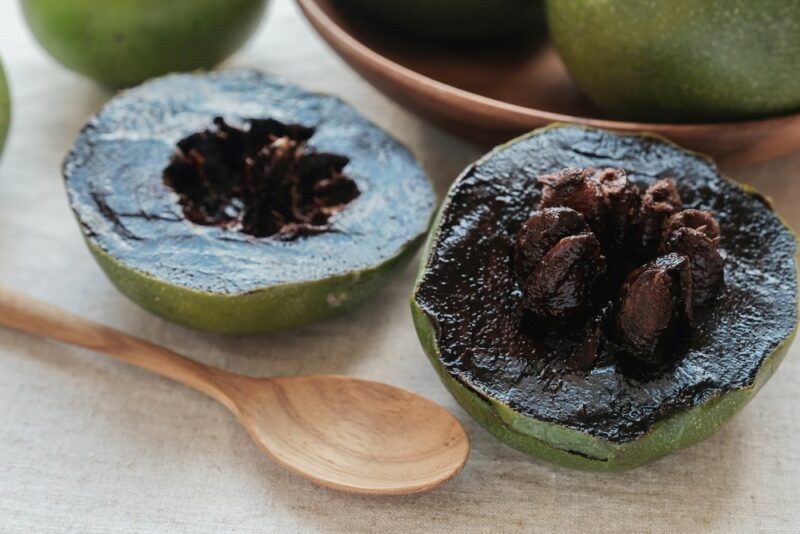 A single black sapote fruit that's been cut in half, showing the dark brown flesh, next to a wooden spoon