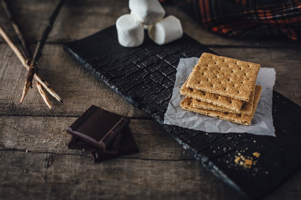 A black slate with the ingredients to make s'mores, including chocolate, marshmallows, graham crackers, and sticks