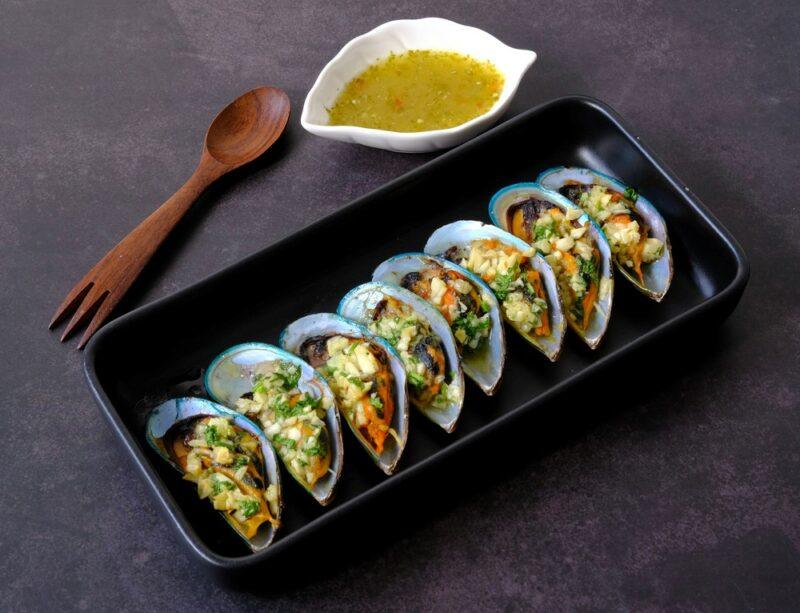 A black dish containing a line of stuffed oysters, next to a spoon/fork and a dish of oil