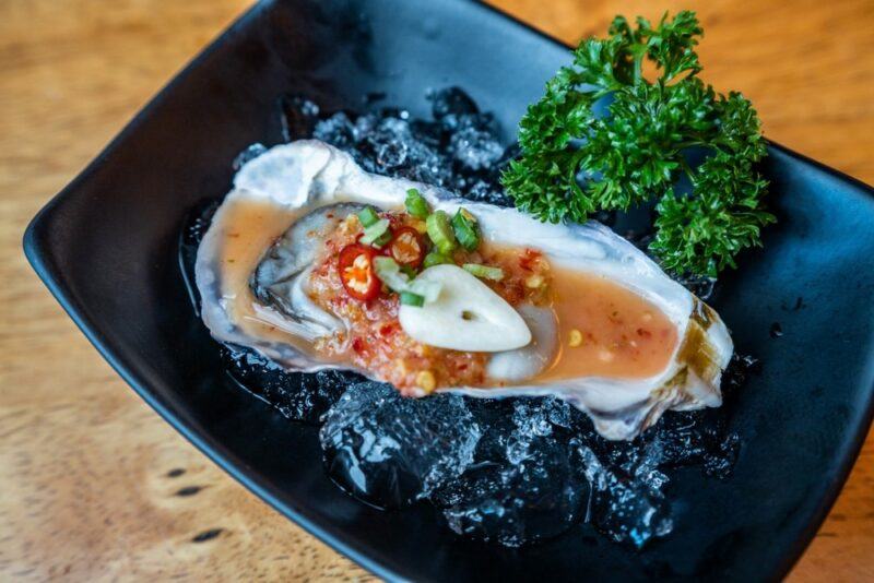 A black plate with a single cooked oyster next to some parsley and ice