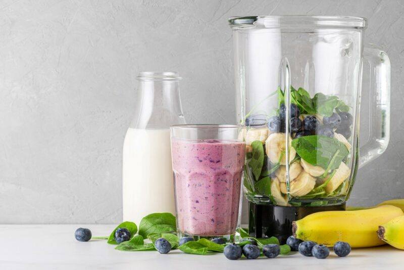 A table with smoothie ingredients, a bottle of milk, a glass containing a berry smoothie, and a blender full of smoothie ingredients