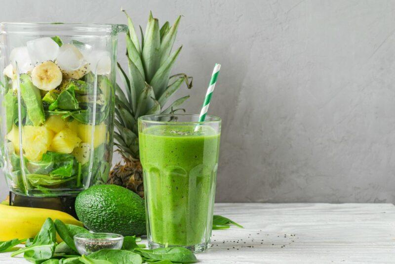 A blender filled with ingredients next to some fruit and vegetables and a glass containing a green smoothie