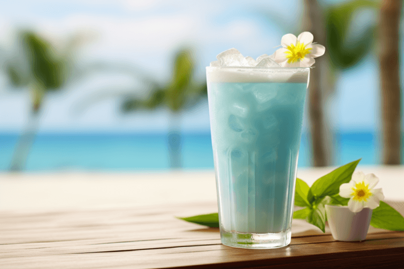 A table with a single blue Hawaiian cocktail, garnished with a flower, with a beach and palm trees in the background