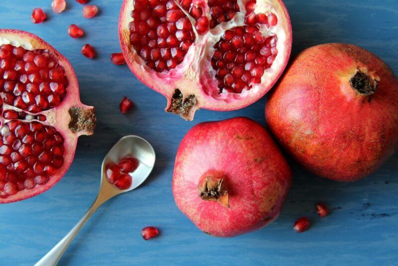 A blue table with two whole pomegranates, two pomegranate halves, and a spoon, with pomegranate arils scattered around