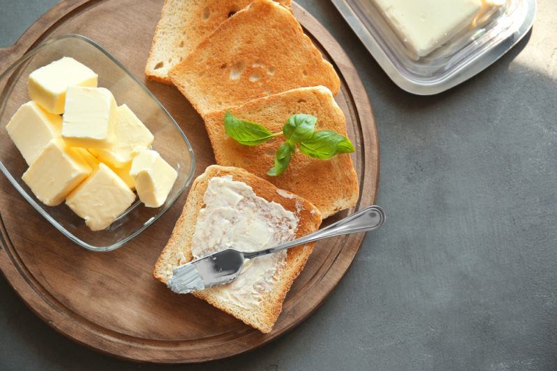 A wooden tray with slices of toast, one that's spread with butter, next to a glass container of butter cubes