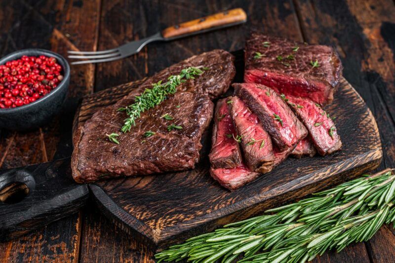 A wooden board with a sliced piece of skirt steak that is garnished with rosemary, next to more rosemary, a fork, and a bowl of red peppercorns