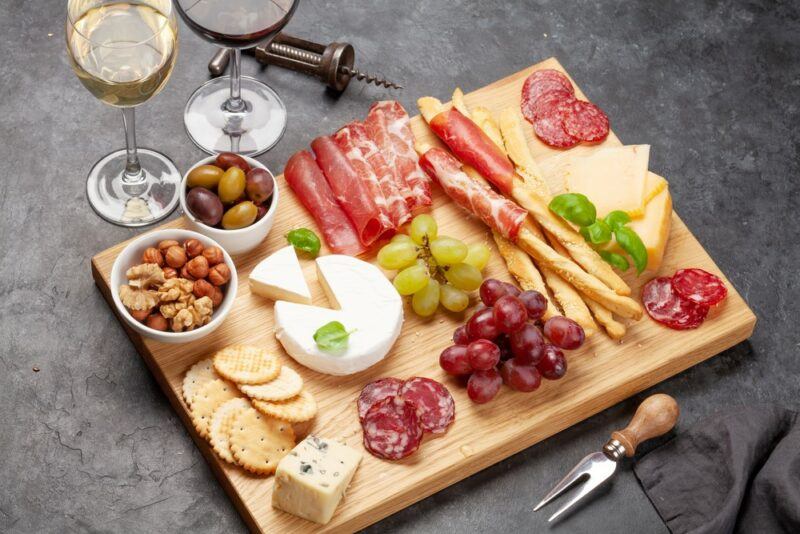 A wooden board with meat, cheese, crackers, and fruit, with a couple of glasses of wine behind it