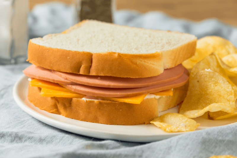 A freshly made bologna sandwich on white bread, next to chips.