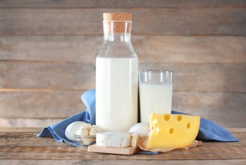 A glass of milk and a bottle of milk on a wooden table, with a blue cloth, some eggs, and three different types of cheese