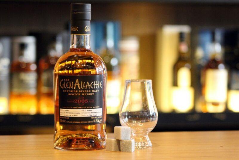 A bottle of GlenAllachie whisky, next to a whisky glass, with out of focus bottles in the background