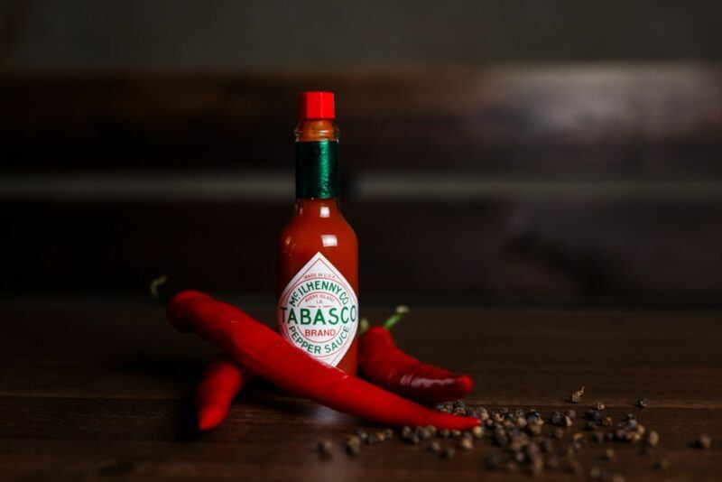 A wooden table with three red chili peppers, tabasco sauce and peppercorns