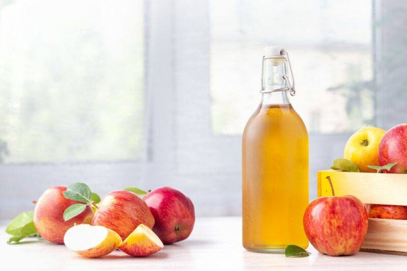 A bottle of apple cider on a kitchen table, near various fresh red apples