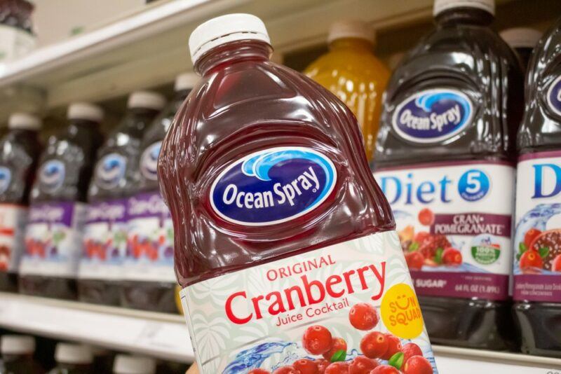 A bottle of cranberry juice cocktail being held up in front of a grocery store shelf filled with other bottles of cranberry juice