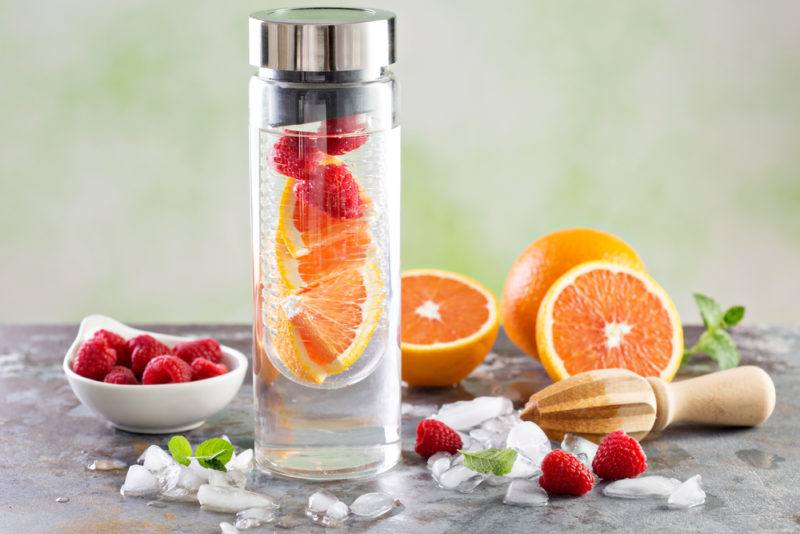 A drink bottle of infused water with oranges, raspberry and a juicer on a table