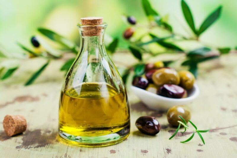 A glass jar of olive oil in front of some olive leaves and a small dish of olives