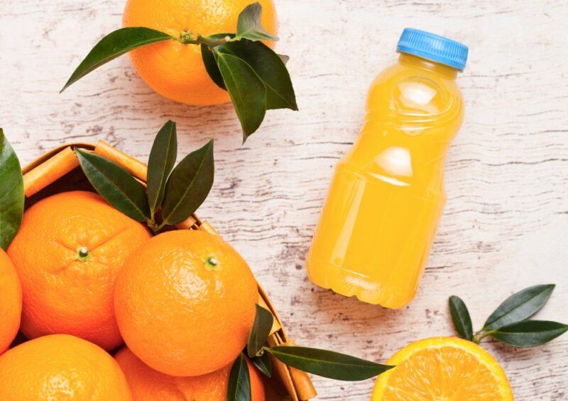 A table with a small bottle of orange juice, with a blue lid, next to a basket of fresh oranges