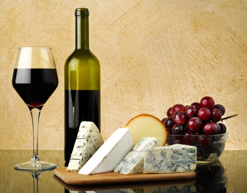 A glass of red wine and a bottle of the wine next to a small board with a selection of cheese and some red grapes