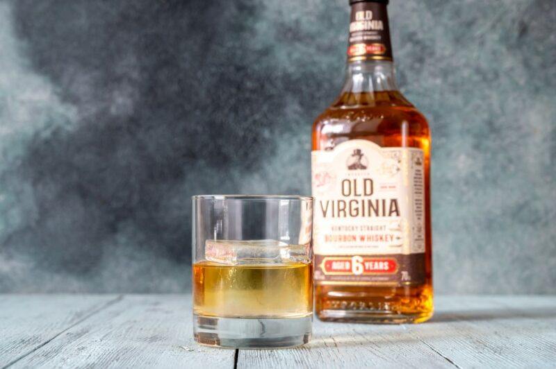 A bottle of bourbon on a table next to a glass of bourbon with a large piece of ice